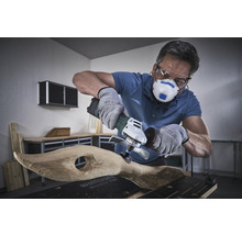 Používateľ s respirátorom, ochrannými okuliarmi a rukavicami opracováva drevo uhlovou brúskou