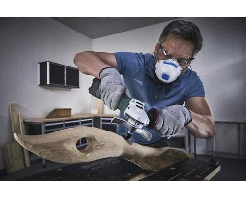 Používateľ s respirátorom, ochrannými okuliarmi a rukavicami opracováva drevo uhlovou brúskou