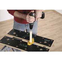 Osoba vŕta vŕtačkou do kusu dreva, ktorý je upevnený na pracovnom stole so žltým upevňovacím prvkom.