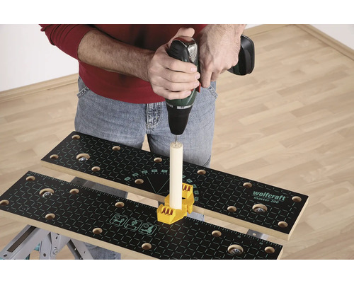 Osoba vŕta vŕtačkou do kusu dreva, ktorý je upevnený na pracovnom stole so žltým upevňovacím prvkom.