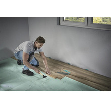 Muž pokladá laminátovú podlahu s kladivom a tĺčikom na kročajovú izoláciu v miestnosti.