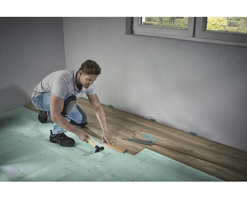 Muž pokladá laminátovú podlahu s kladivom a tĺčikom na kročajovú izoláciu v miestnosti.