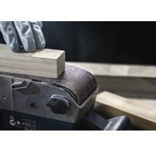 Detailný záber pásovej brúsky brúsiacej kus dreva, obsluhovaný osobou v rukaviciach.