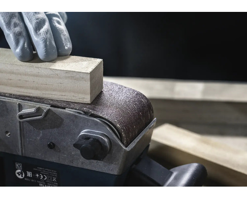 Detailný záber pásovej brúsky brúsiacej kus dreva, obsluhovaný osobou v rukaviciach.