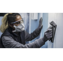 Osoba nosí ochranné pomôcky a používa brúsnu dosku na brúsenie steny.