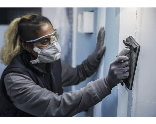 Osoba nosí ochranné pomôcky a hladí stenu pomocou hladidla.