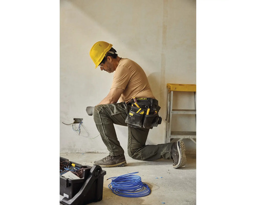 Elektrikár kľačí a pracuje na elektrických vodičoch s opaskom na náradie a žltou ochrannou helmou.