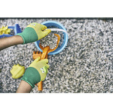 Ruky v záhradných rukaviciach plnia vedro lopatkou a kameňmi, obklopené hračkami do pieskoviska.