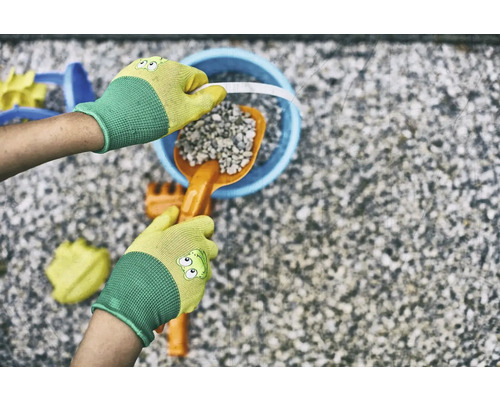 Ruky v záhradných rukaviciach plnia vedro lopatkou a kameňmi, obklopené hračkami do pieskoviska.