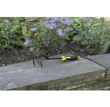 Záhradný kultivátor s tromi hrotmi a plastovou rukoväťou pred kvetinovým záhonom