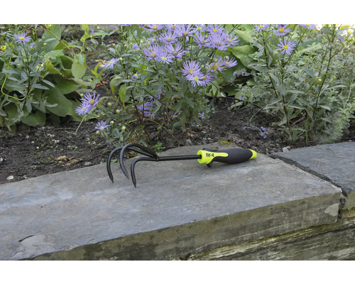 Záhradný kultivátor s tromi hrotmi a plastovou rukoväťou pred kvetinovým záhonom