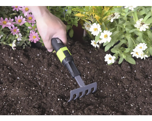 Ručné hrabličky s čierno-zelenou rukoväťou a for_q Logo používané v tmavej zemine medzi ružovými a bielymi kvetmi.