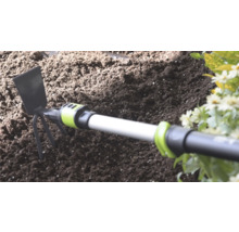 Ručná motyčka s kovovou čepeľou a tromi hrotmi v tmavej záhradnej zemine vedľa kvetov.