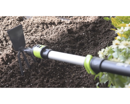 Ručná motyčka s kovovou čepeľou a tromi hrotmi v tmavej záhradnej zemine vedľa kvetov.