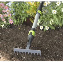 Malé záhradné hrable so sivou kovovou hlavou a nastaviteľnou teleskopickou násadou v zemine pred kvetmi.