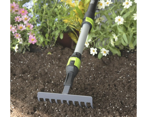 Malé záhradné hrable so sivou kovovou hlavou a nastaviteľnou teleskopickou násadou v zemine pred kvetmi.