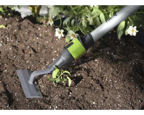 Záhradná motyka so sivou čepeľou a zelenou spojkou pri pletí buriny v tmavej pôde vedľa rastliny.