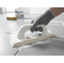 Osoba v ochrannej rukavici nanáša pomocou epoxidového hladidla bielu škárovaciu hmotu na sivú dlažbu.