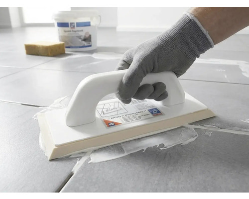 Osoba v ochrannej rukavici nanáša pomocou epoxidového hladidla bielu škárovaciu hmotu na sivú dlažbu.