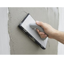 Ruka vyhladzuje čerstvú omietku na stene pomocou hubového hladidla.