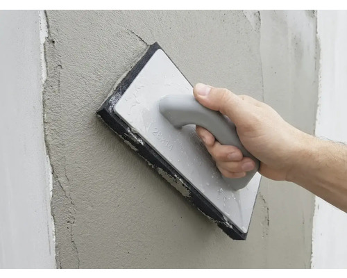 Ruka vyhladzuje čerstvú omietku na stene pomocou hubového hladidla.