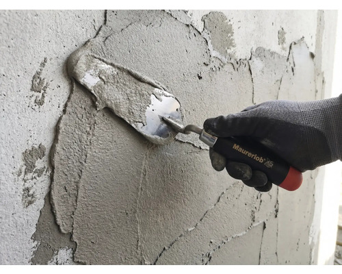 Ruka v pracovnej rukavici nanáša maltu na stenu pomocou murárskej lyžice Maurerlob.