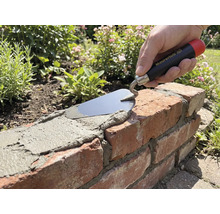 Ruka s murárskou lyžicou Maurerlob nanáša maltu na tehlový múrik v záhrade.