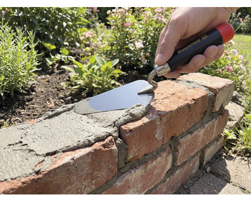 Ruka s murárskou lyžicou Maurerlob nanáša maltu na tehlový múrik v záhrade.