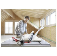 Muž s ochrannými okuliarmi reže drevenú dosku stolovou kotúčovou pílou vo svetlej miestnosti s drevenými trámami a OSB doskami.