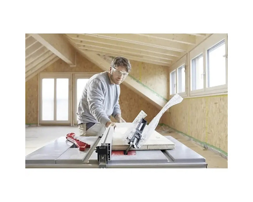 Muž s ochrannými okuliarmi reže drevenú dosku stolovou kotúčovou pílou vo svetlej miestnosti s drevenými trámami a OSB doskami.