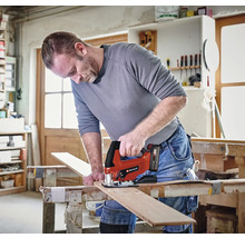 Remeselník v jasnej dielni reže drevenú dosku na pracovnom stole pomocou akumulátorovej priamočiarej píly Einhell. Einhell Logo.