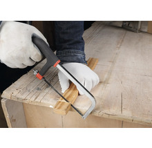 Osoba v bielych ochranných rukaviciach reže drevenú lištu malou ručnou pílkou na pracovnom stole.