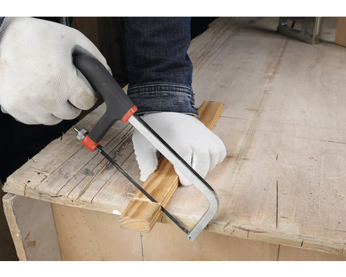 Osoba v bielych ochranných rukaviciach reže drevenú lištu malou ručnou pílkou na pracovnom stole.