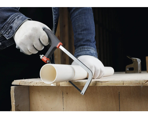 Osoba v rukaviciach reže bielu plastovú rúru malou pílkou na kov na drevenom pracovnom stole.