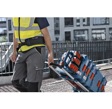 Remeselník v reflexnej veste ťahá transportný vozík naložený kuframi na náradie Bosch L-BOXX a stavebným rádiom na stavenisku.