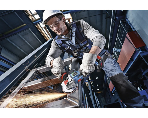 Remeselník v ochrannom odeve opracováva oceľovú konštrukciu akumulátorovou uhlovou brúskou, od ktorej lietajú iskry.