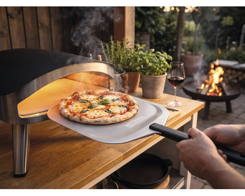 Čerstvo upečená pizza sa vyberá z prenosnej pece na pizzu na drevenom stole v záhrade pomocou lopaty na pizzu s Tenneker Logo.