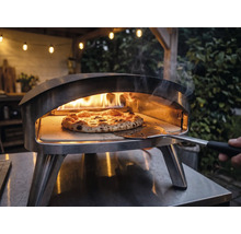 Vonkajšia nerezová pec na pizzu s horiacimi plameňmi a pizzou na pizzovom kameni, s ktorou sa manipuluje pomocou lopaty na pizzu.