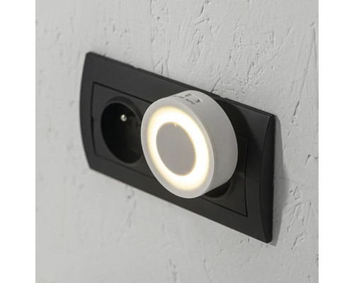 Okrúhle biele nočné LED svetlo so senzorom do zásuvky, zapojené do nástennej zásuvky.