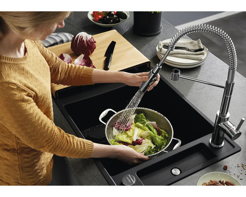 Osoba umýva šalát v kovovom cedníku v čiernom dreze s vyťahovacou kuchynskou batériou a pružinou v modernej kuchyni.