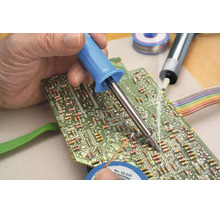 Ruka prispájkuje elektronickú súčiastku na zelenú dosku plošných spojov pomocou spájkovačky.