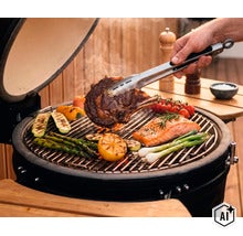 Ruka obracia steak pomocou grilovacích klieští na keramickom grile so zeleninou, lososom a paradajkami na grilovacom rošte. (Image AI modified or generated)