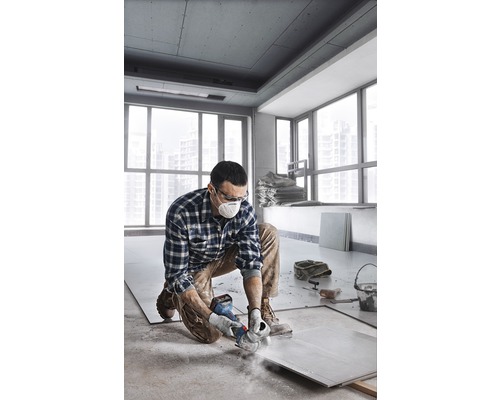 Muž kľačí v miestnosti a reže dlaždicu brúskou. Má na sebe ochrannú masku a ochranné okuliare.