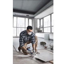 Muž kľačí v miestnosti a reže dlaždicu uhlovou brúskou. Má na sebe ochranný odev.