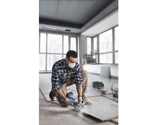 Muž kľačí v miestnosti a reže dlaždicu uhlovou brúskou. Má na sebe ochranný odev.