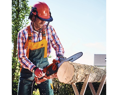 Muž pília elektrickou reťazovou pílou klát na kozlíku a má na sebe helmu s tvárovým štítom a chrániče sluchu