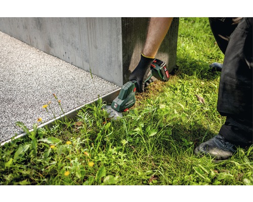 Záhradník strihá trávu akumulátorovými nožnicami na trávu značky Metabo pozdĺž betónového múru.