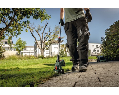 Osoba používa akumulátorový vyžínač okrajov trávnika v záhrade, v pozadí je vidieť kosačku na trávu.