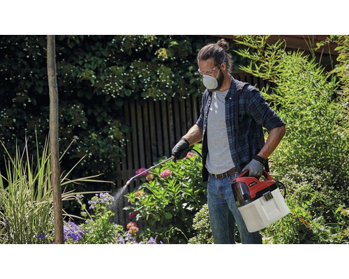 Muž s ochrannými okuliarmi a respirátorom používa akumulátorový postrekovač rastlín v záhrade.