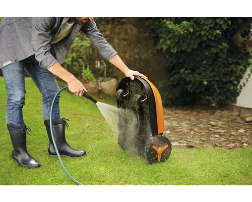 Muž čistí robotickú kosačku na trávu pomocou hadice na trávniku.